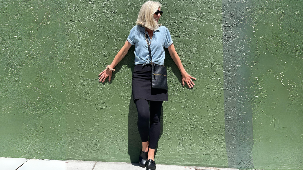 Woman in blue shirt and black skirted leggings leaning against green wall, showcasing Fashion Over 50 Athleisure.