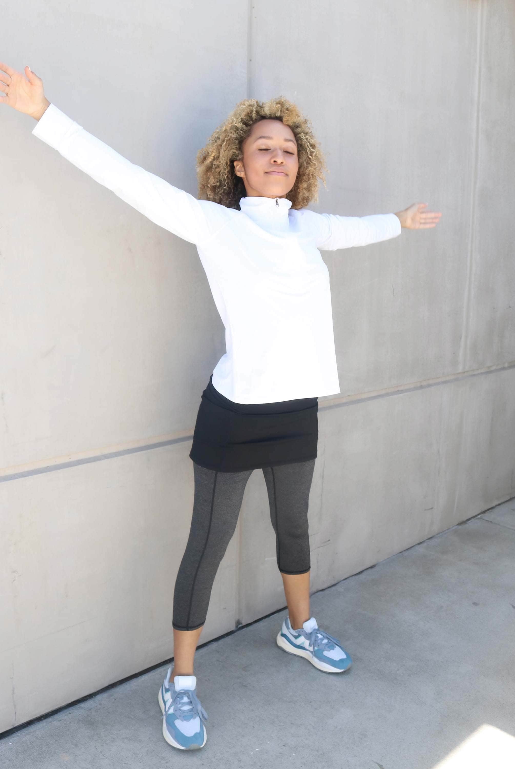 One piece black skirt with heather grey capris length leggings. Model is standing near a building with arms spread wide wearing white our a white zip front activewear shirt.