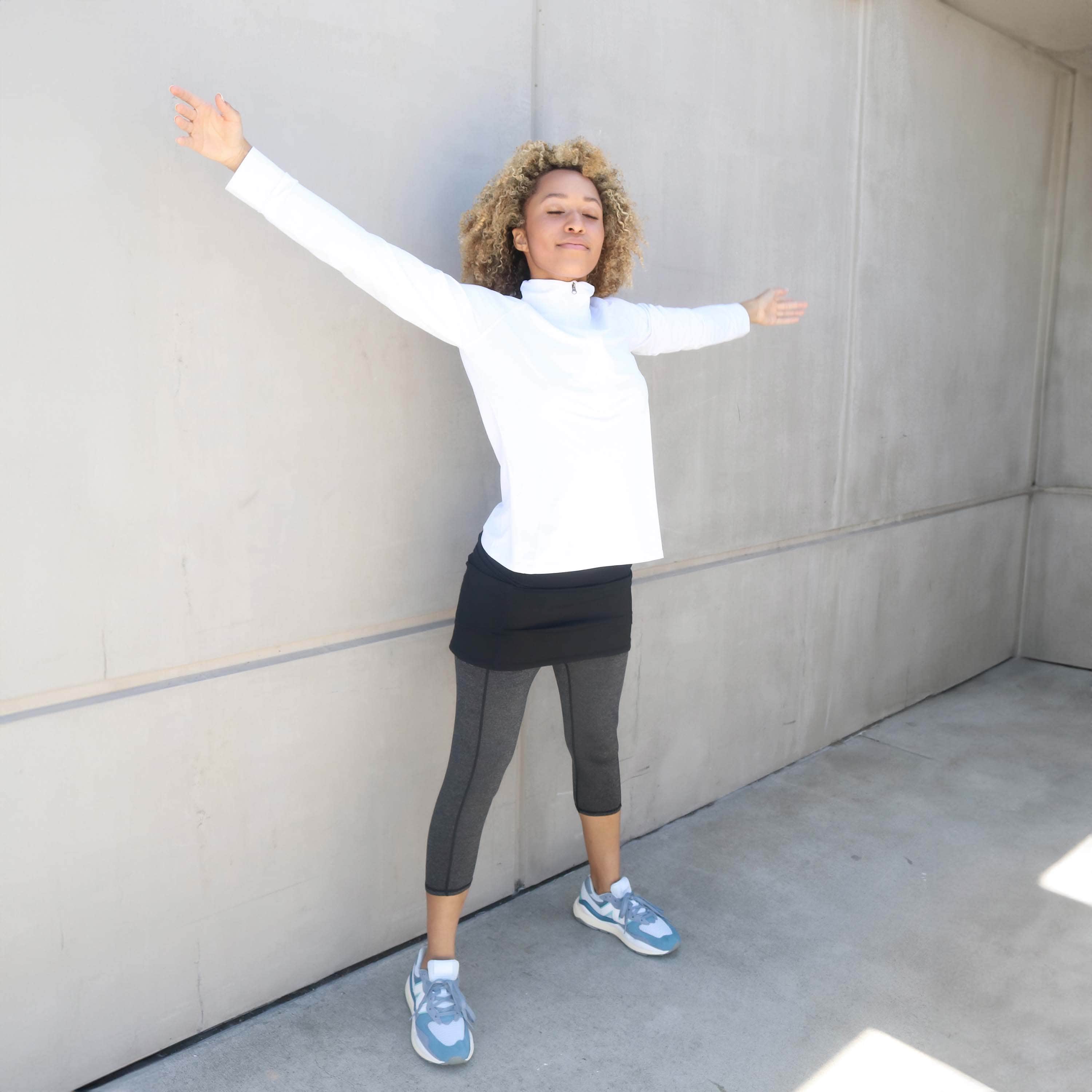 One piece black skirt with heather grey capris length leggings. Model is standing near a building with arms spread wide wearing white our a white zip front activewear shirt.