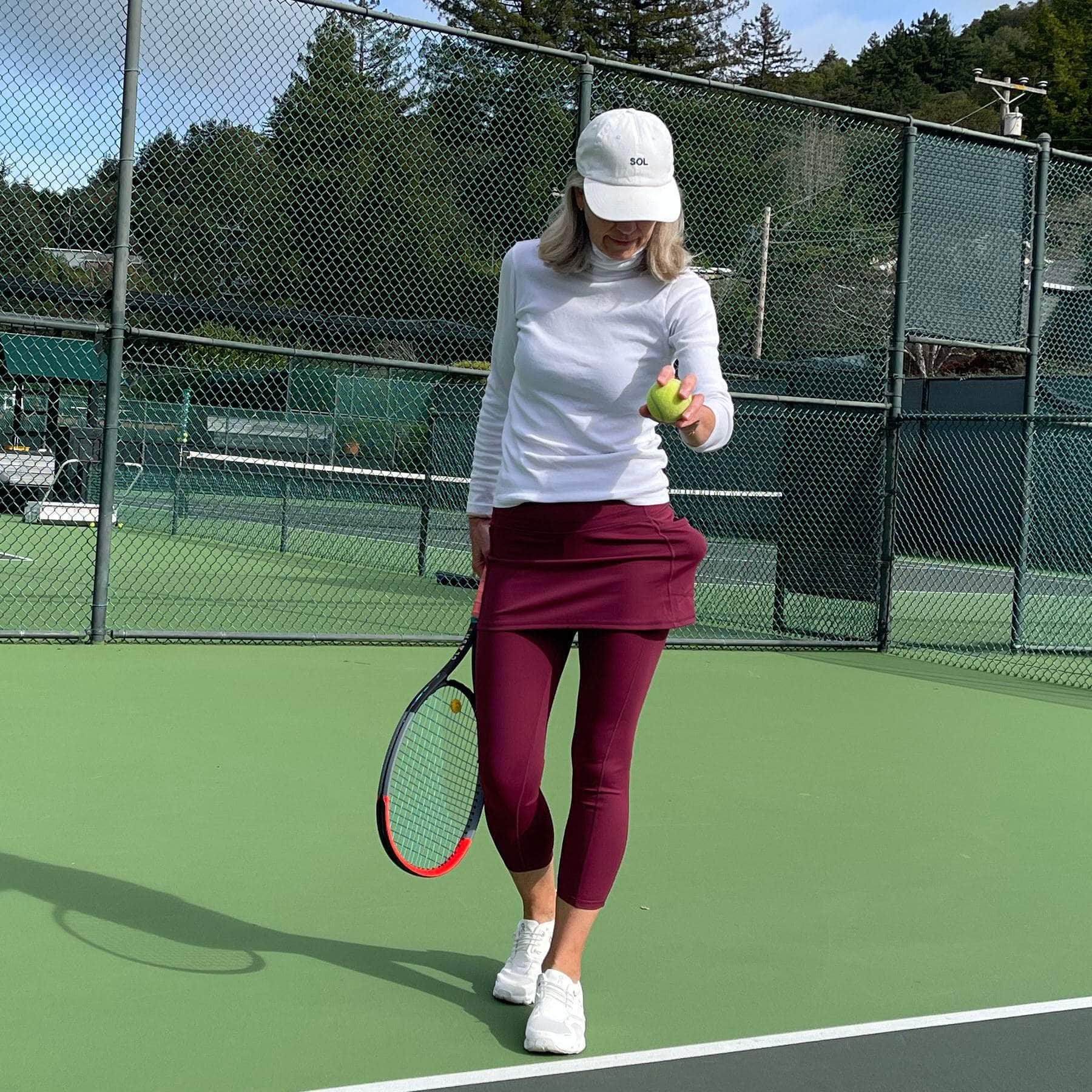 Woman in pretty dark red skirted leggings on tennis court about to serve.