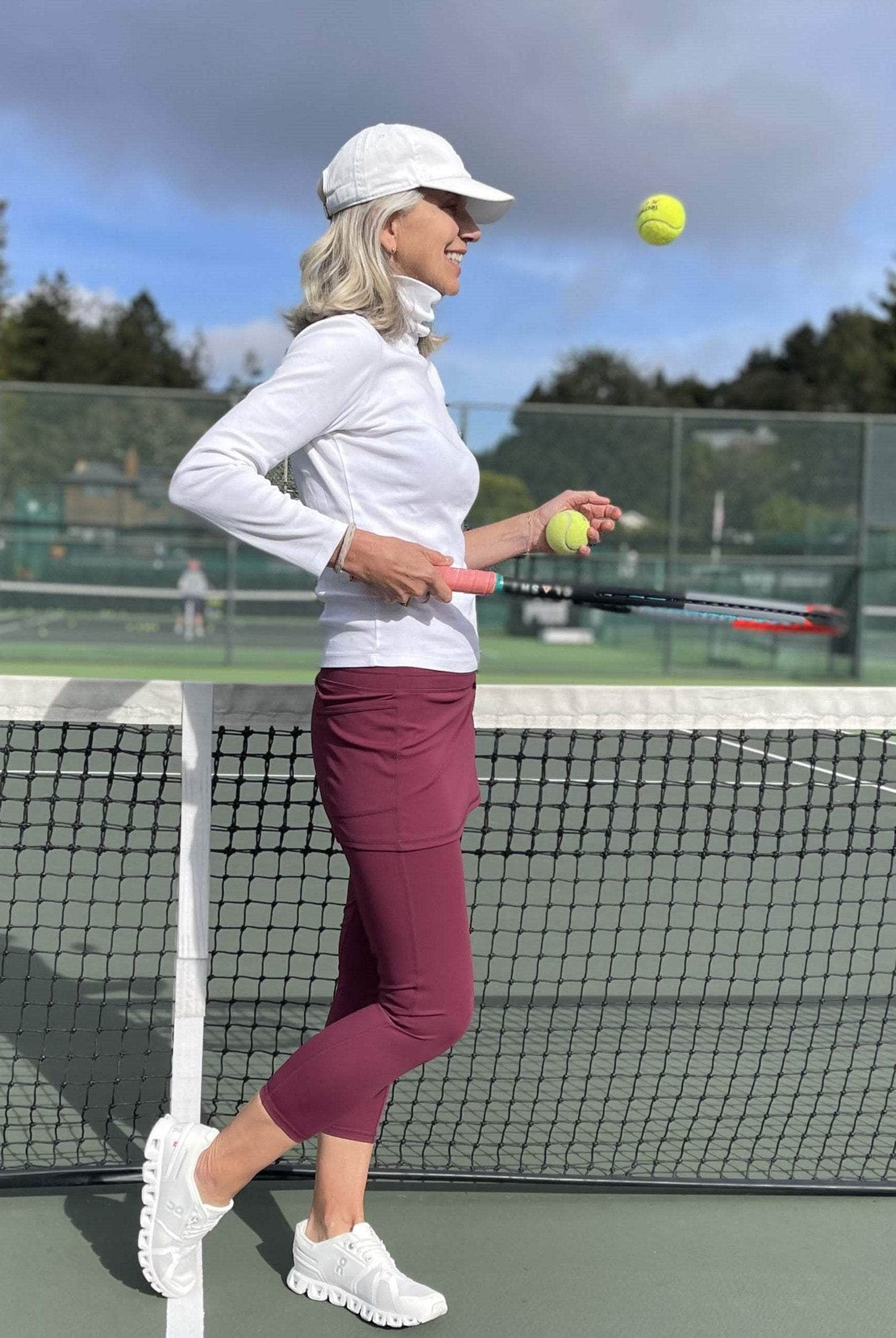 Model on tennis court wearing cranberry colored 2-1 skirted leggings with white turtleneck and white tennis cap.