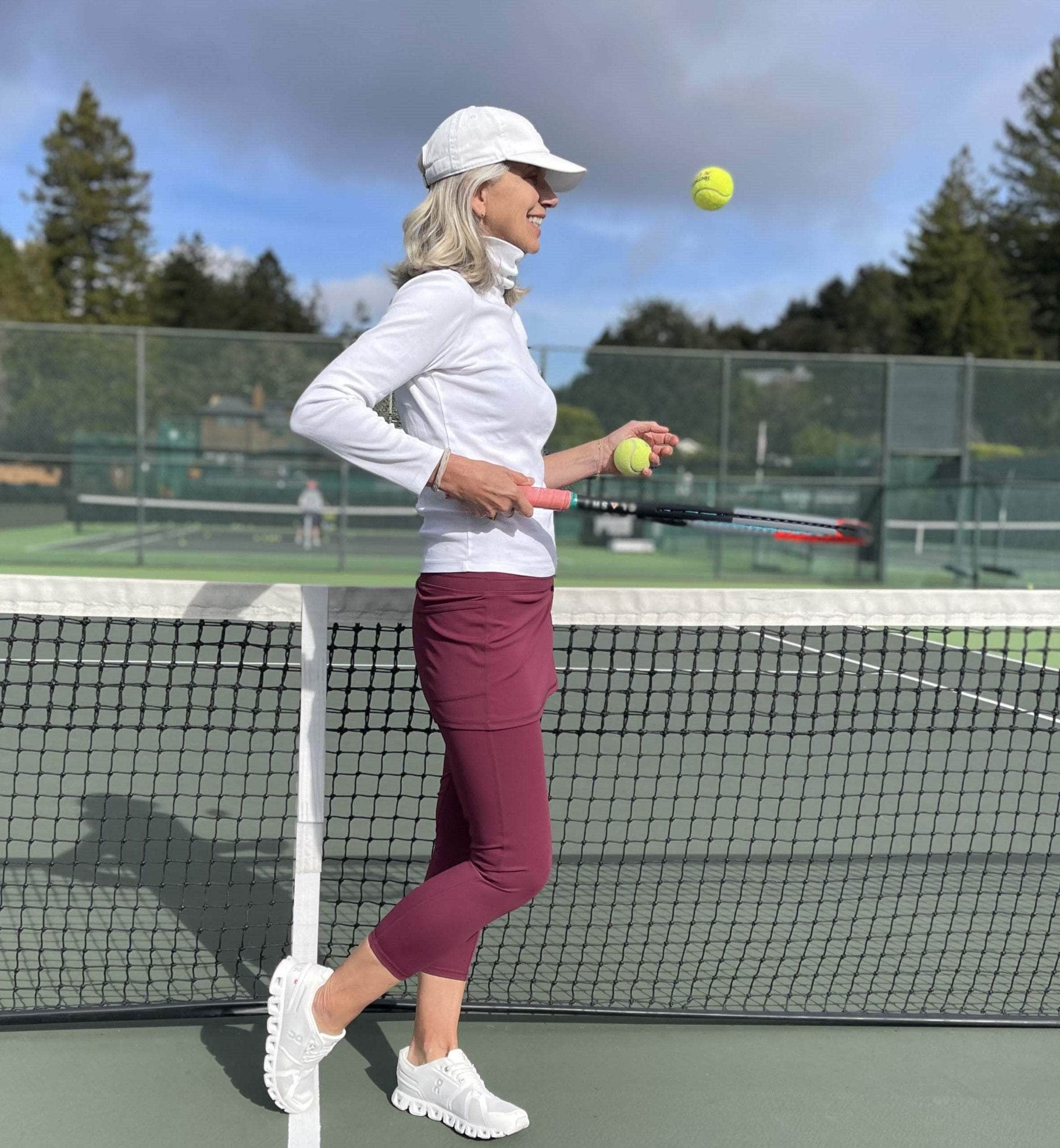 Model on tennis court wearing cranberry colored 2-1 skirted leggings with white turtleneck and white tennis cap.