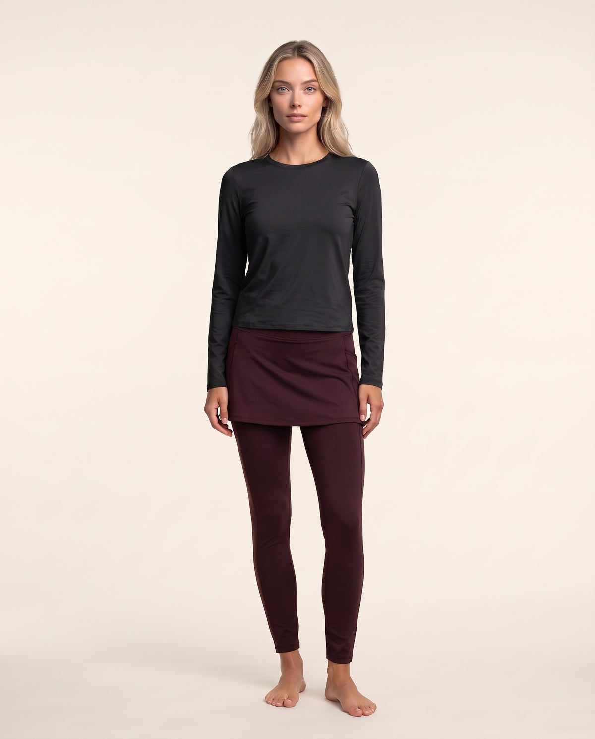 A woman stands barefoot against a plain light beige studio background, wearing a dark charcoal long-sleeve top, a dark burgundy mini skirt, and matching burgundy leggings.