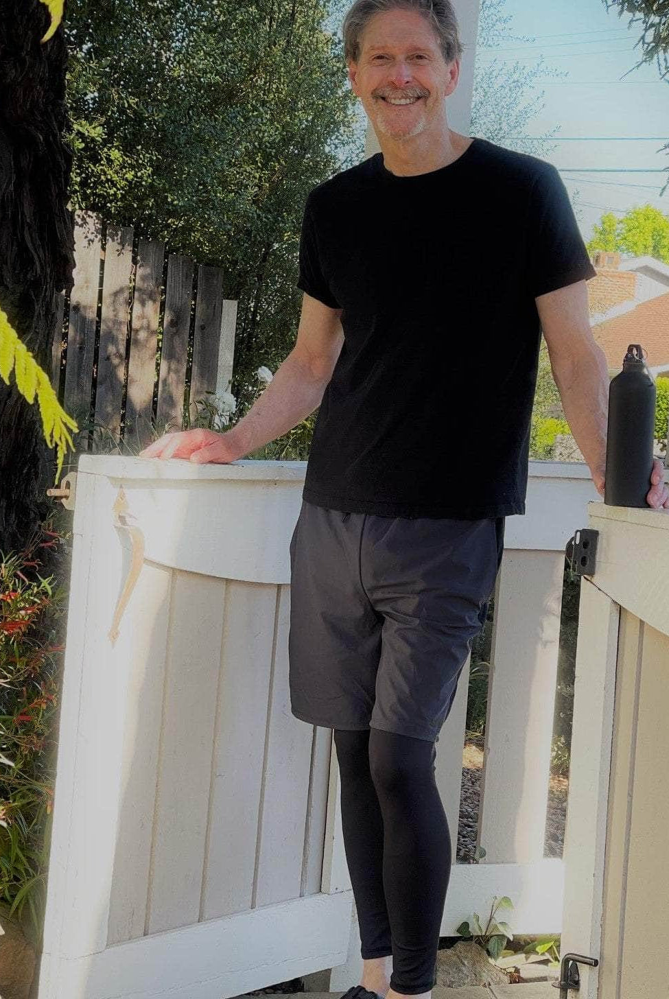 Male model wearing Sol Sister Mister Men's 2-1 Shorts with Leggings with Black T-Shirt. Front view of model standing in front of gate.