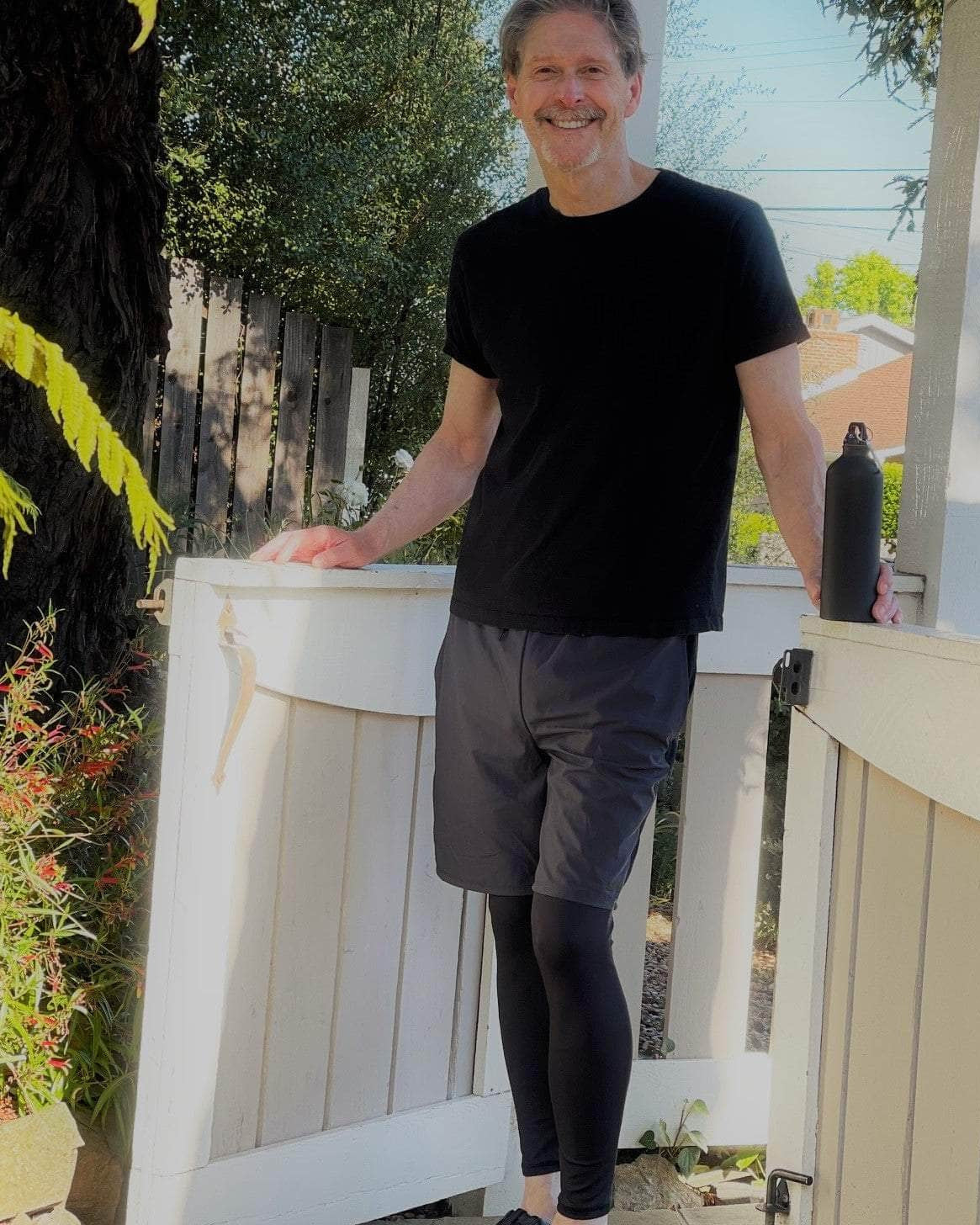 Male model wearing Sol Sister Mister Men's 2-1 Shorts with Leggings with Black T-Shirt. Front view of model standing in front of gate.