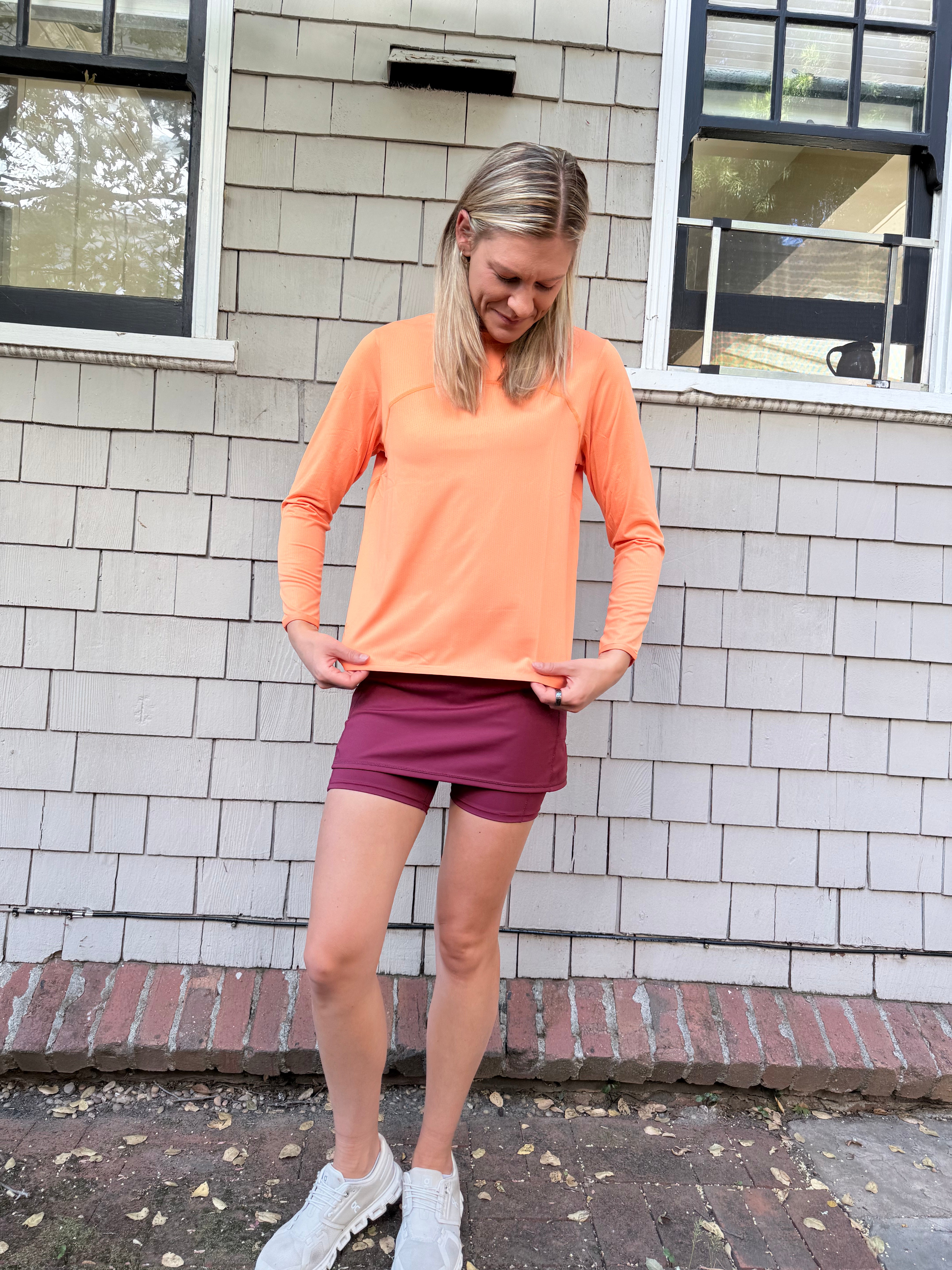 A woman standing near a shingled wall wearing a orange Ventura Mesh Pullover, a red tennis skirt, and white sneakers.