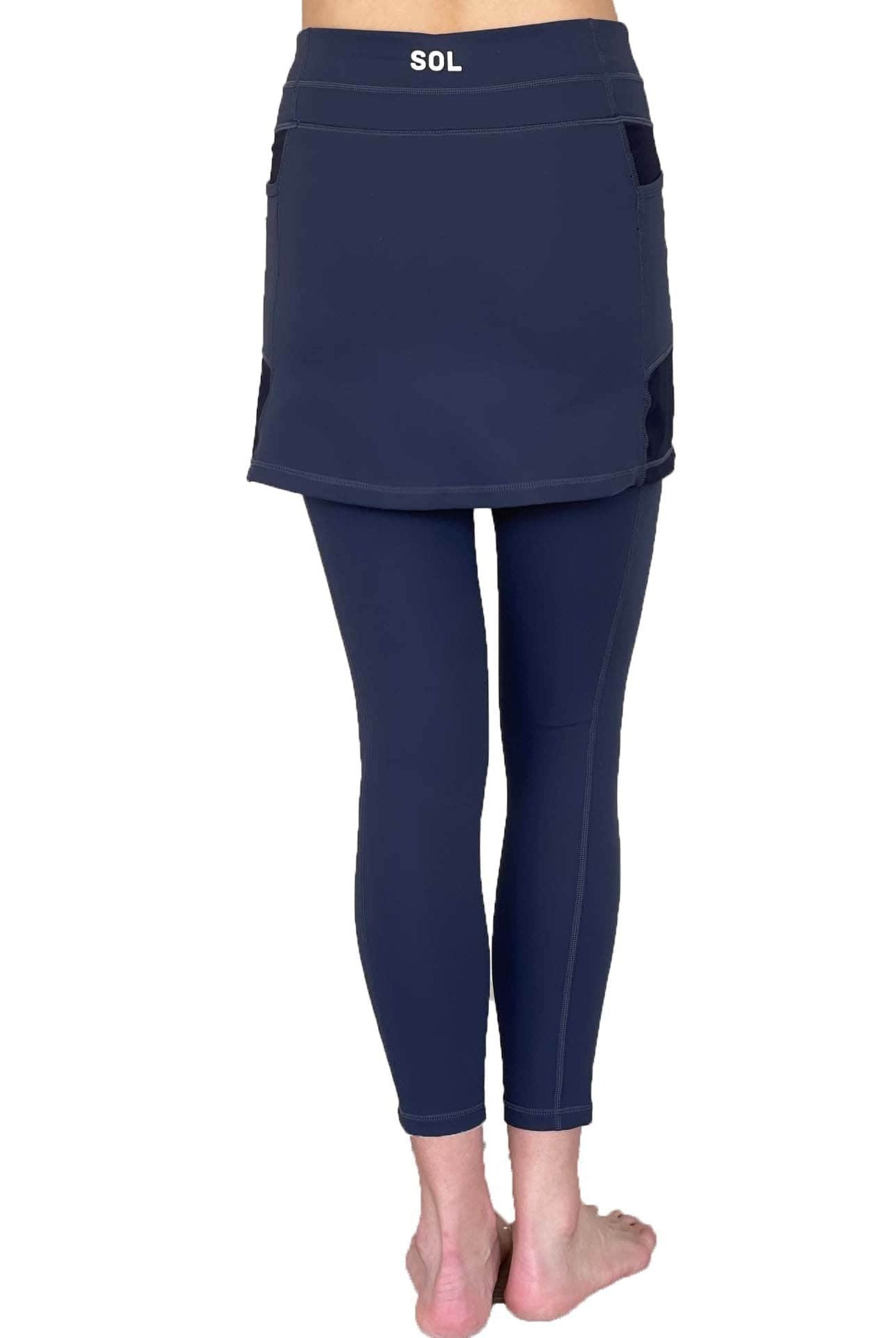 Woman wearing dark blue 7/8th legging length skirted leggings from waist down with white background from back view.
