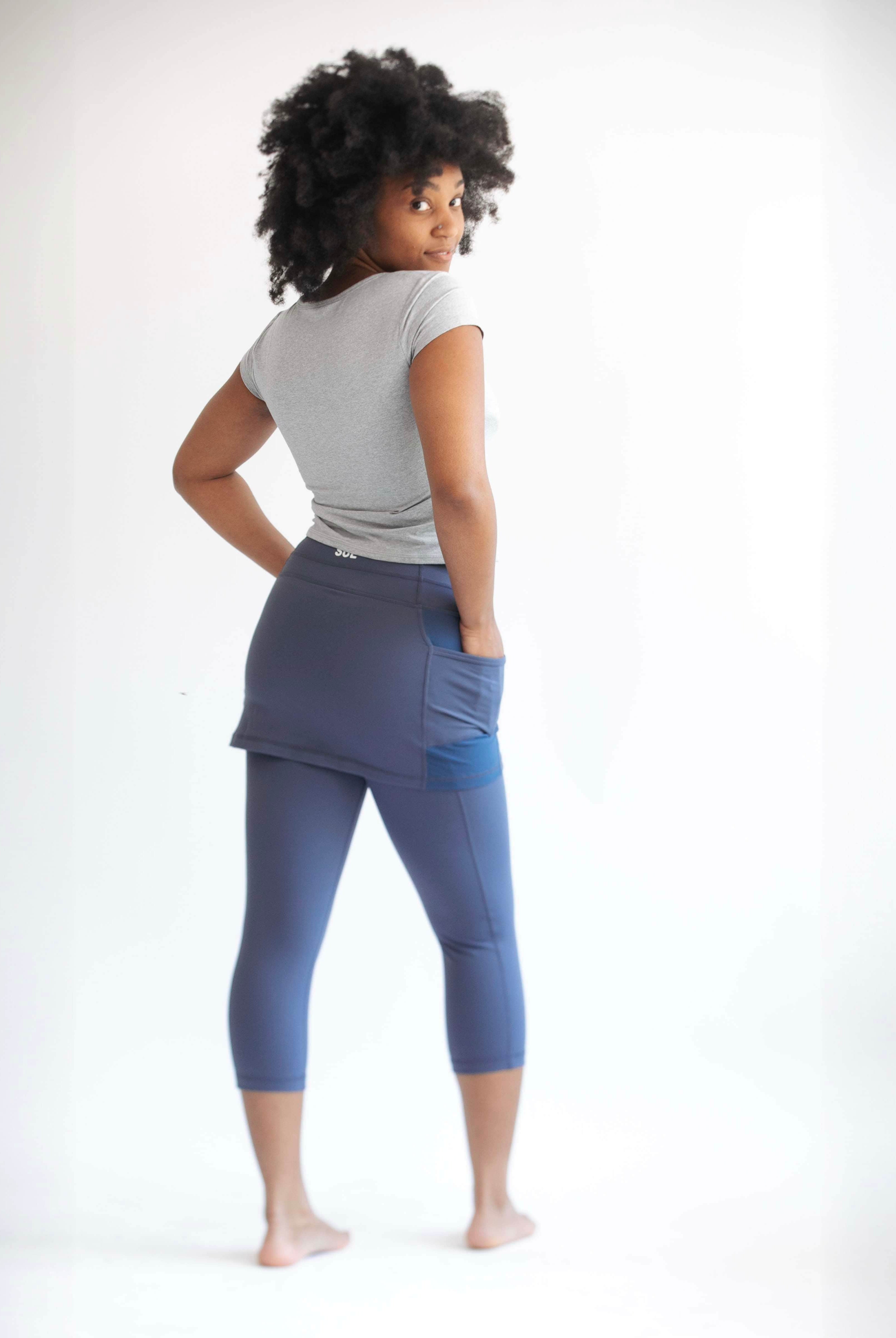 Rear studio shot of model wearing blue grey capris length leggings with attached skirt.