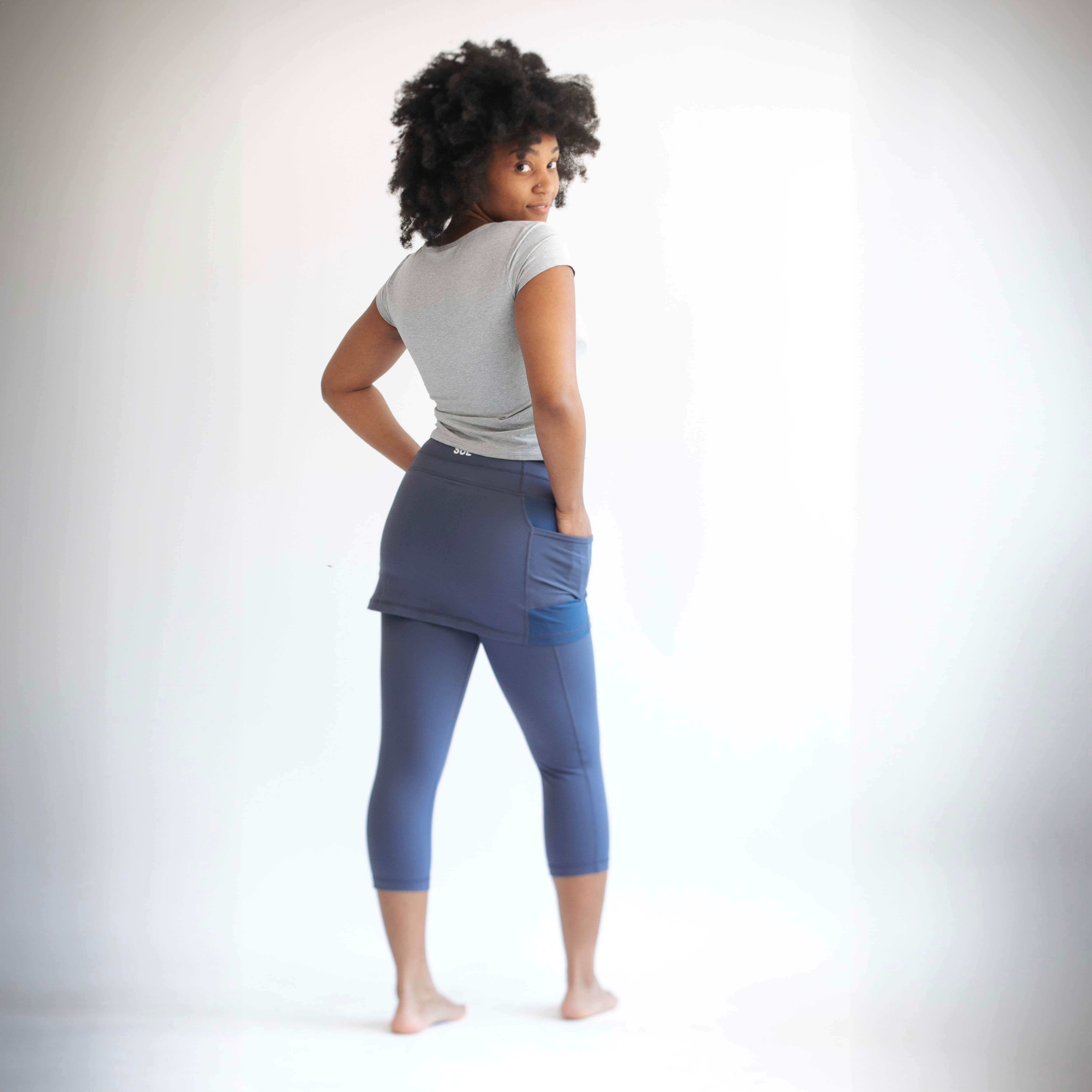 Rear studio shot of model wearing blue grey capris length leggings with attached skirt.