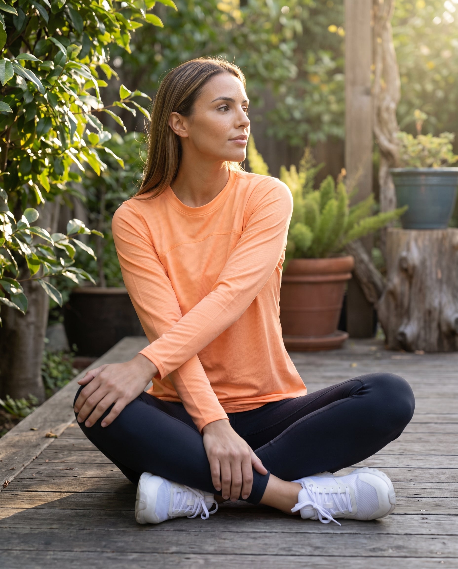 A woman sitting crosslegged on a wooden deck wearing an orange mesh pullover and black leggings