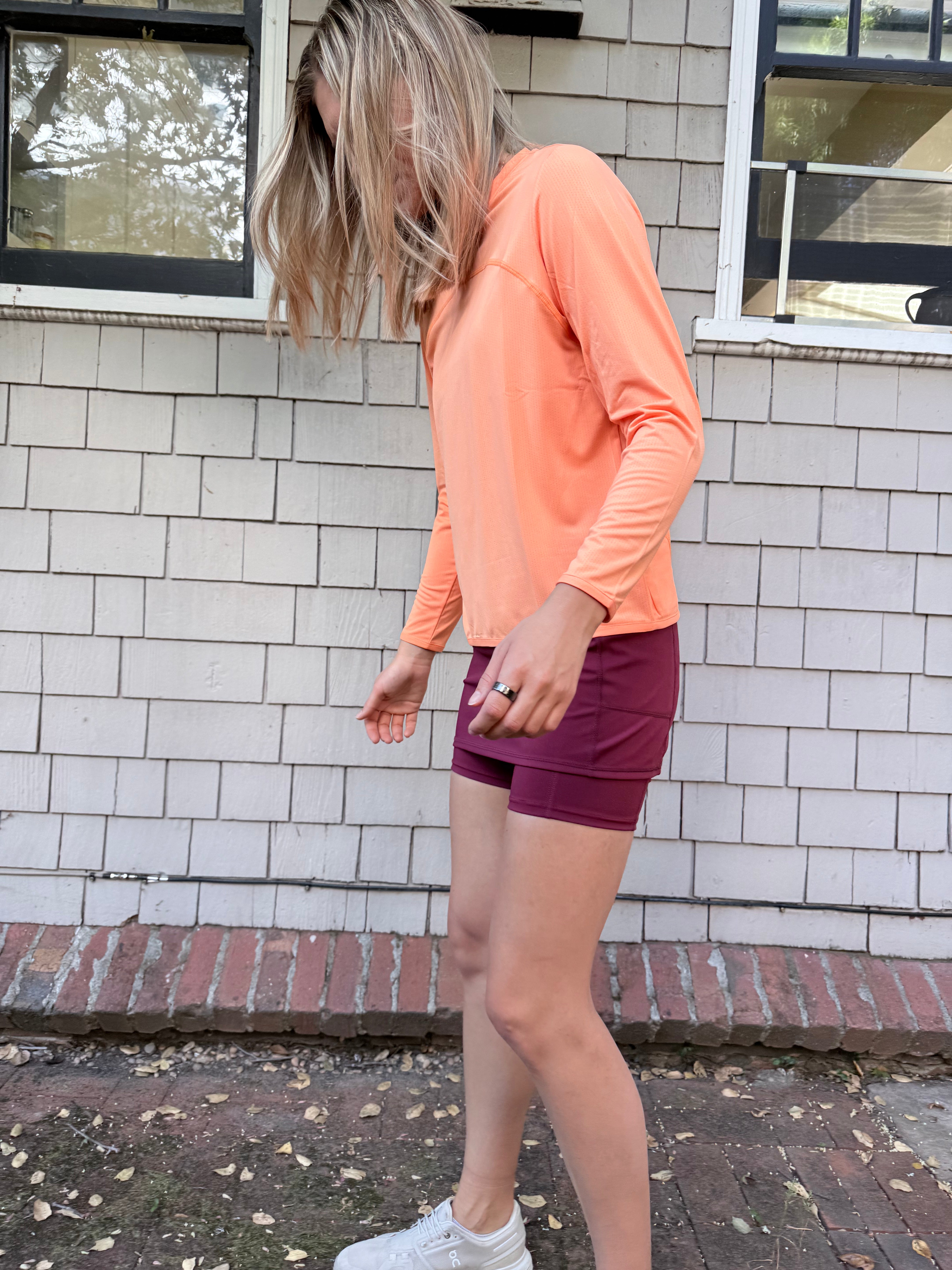 A woman standing near a shingled wall wearing a orange Ventura Mesh Pullover, a red tennis skirt, and white sneakers. The perfect long sleeve UPF sun protective shirt for women.