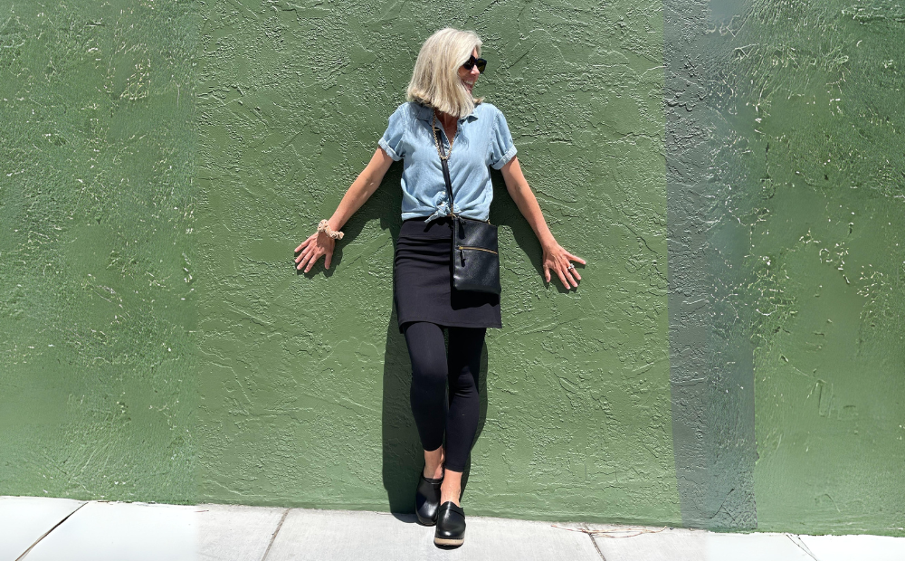Woman in blue shirt and black skirted leggings leaning against green wall, showcasing Fashion Over 50 Athleisure.