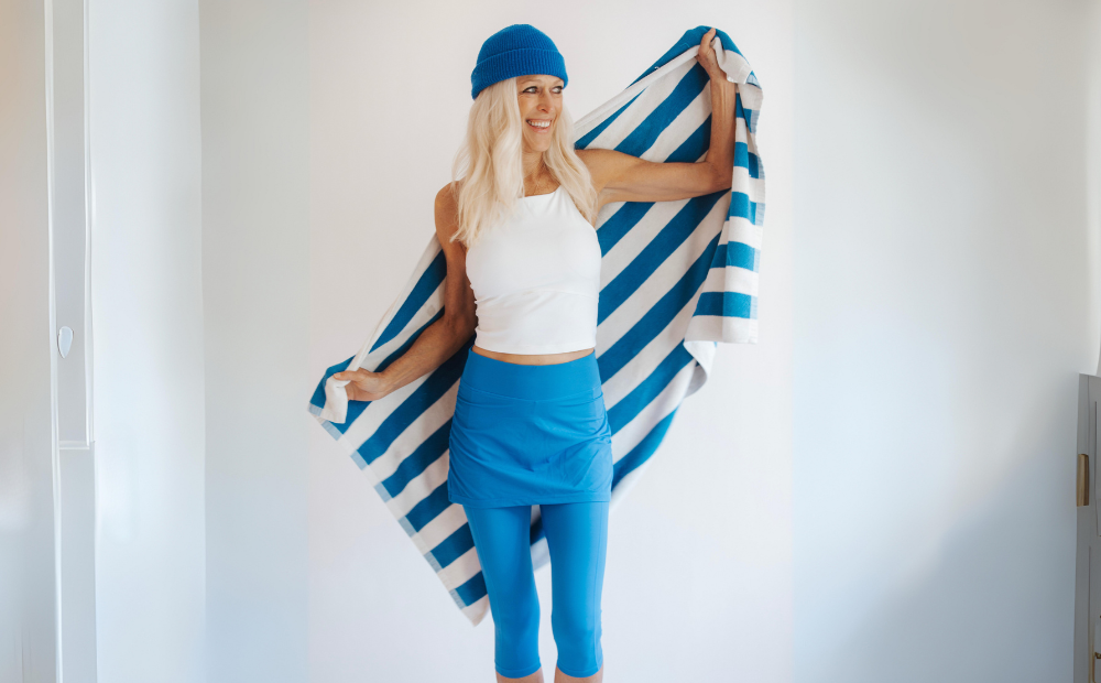 A woman wearing a blue hat and blue leggings holds a towel. She is ready for summer with her essential skirted swim leggings.