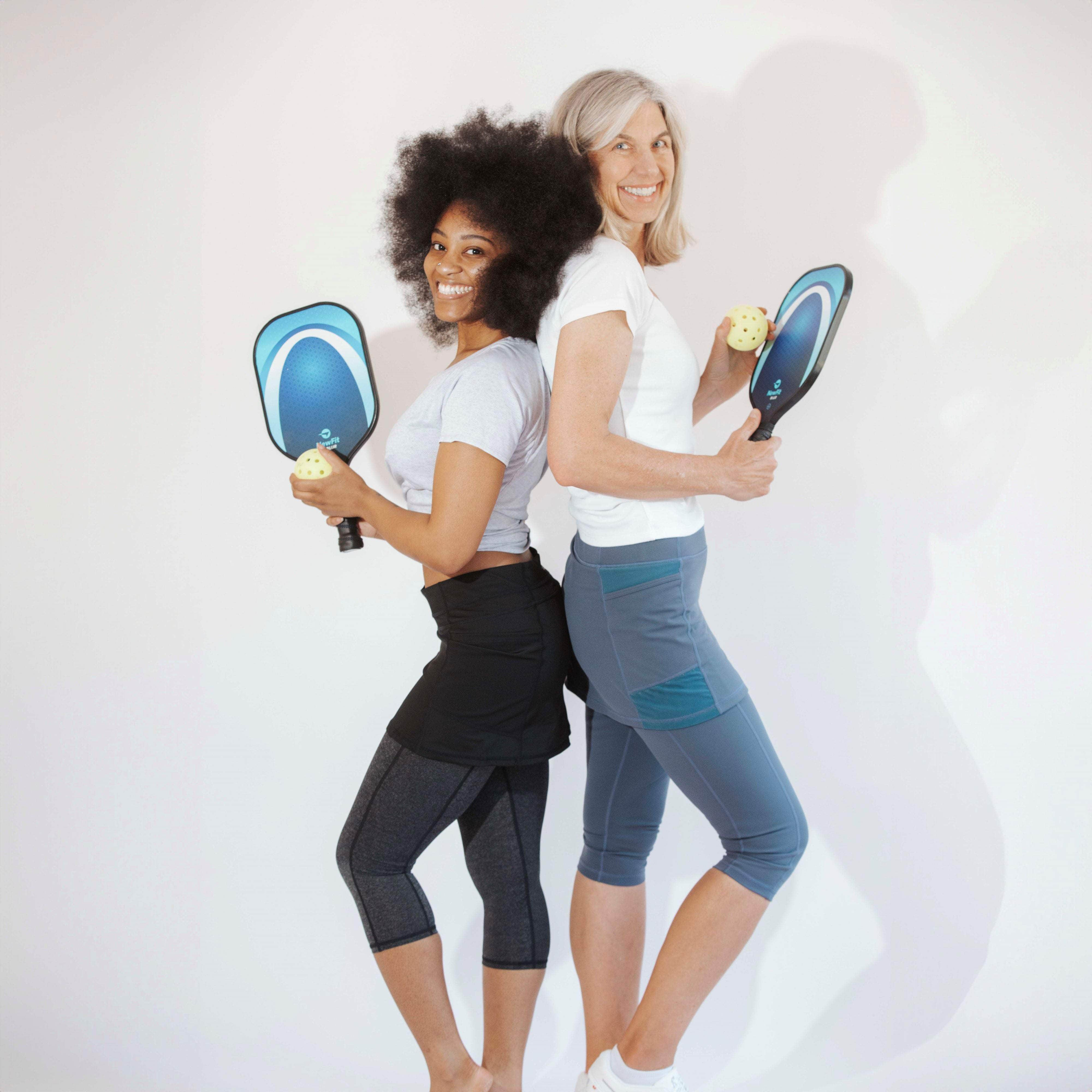 Two models are standing back to back with pickleball paddles.  One is wearing One piece black skirt with heather grey capris length leggings.  Other model is taller and wearing blue grey  skirted capris leggings.  Both have short sleeve activewear shirts