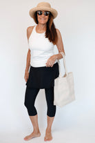Woman wearing a white tank top, black capri pants, straw hat, and sunglasses, holding a white tote bag on a white background.