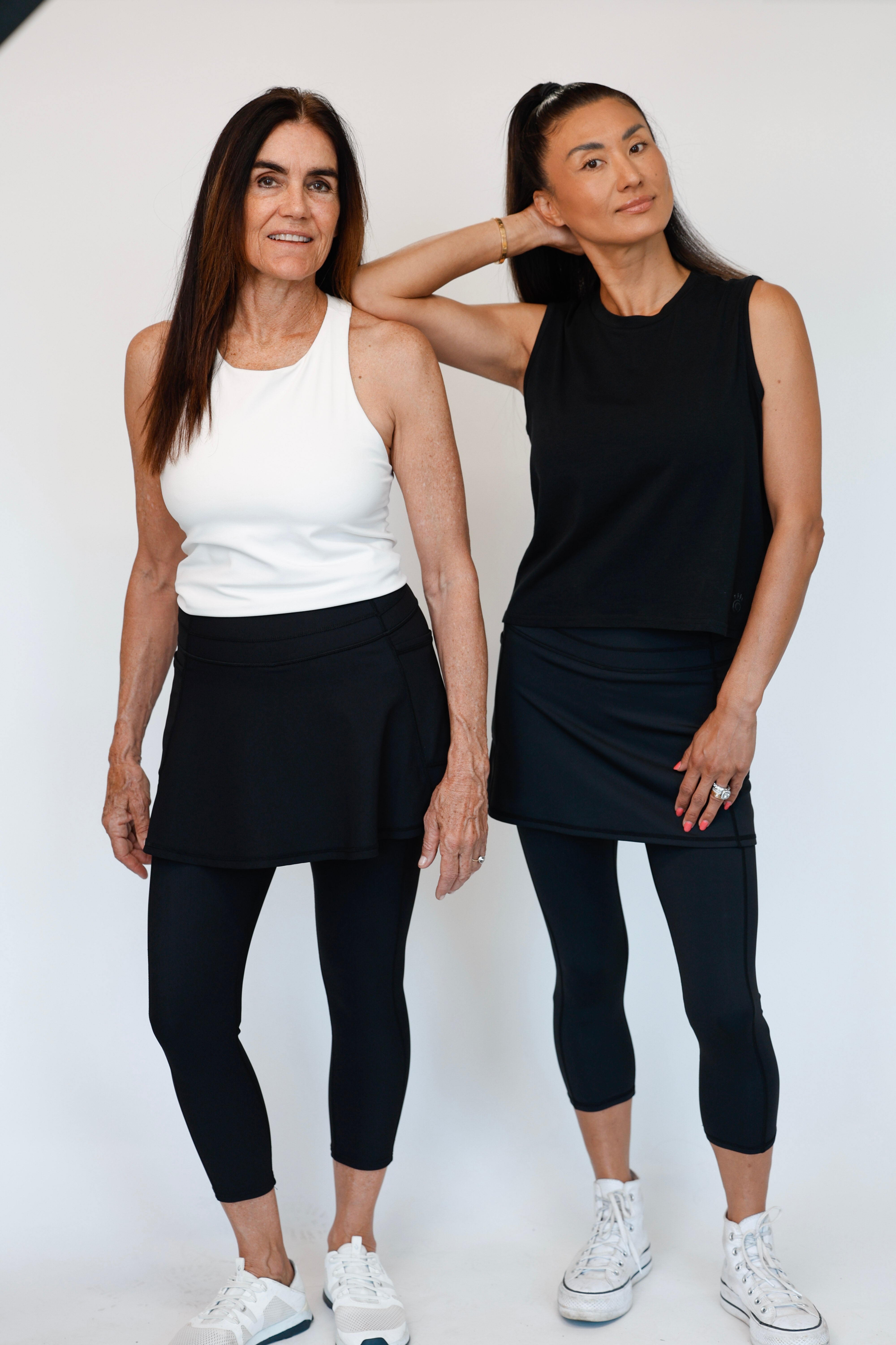 Two women wearing black flare ribbed skirted leggings and white athletic shirts standing against a white background. Comfortable modest feminine active wear built for travel and built for movement; perfect for tennis, pickleball, golf, yoga, pilates and hiking. UPF sun protective and moisture wicking skirted leggings from a California based women founded and owned athletic wear company called Sol Sister Sport. 