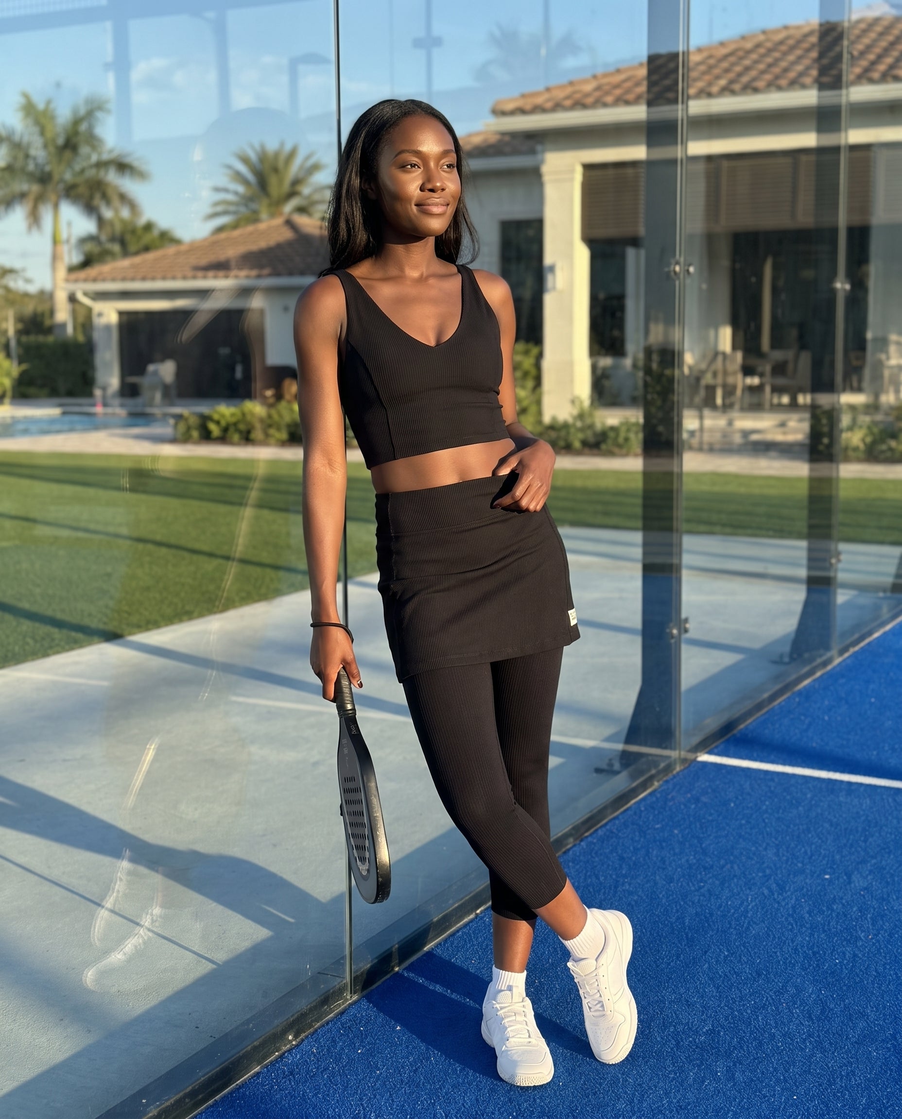 A woman stands on a blue tennis court wearing a matching black athletic set consisting of a crop top, black ribbed skirt with attached leggings, holding a tennis racket. The background shows a glass wall, green grass, and a house.