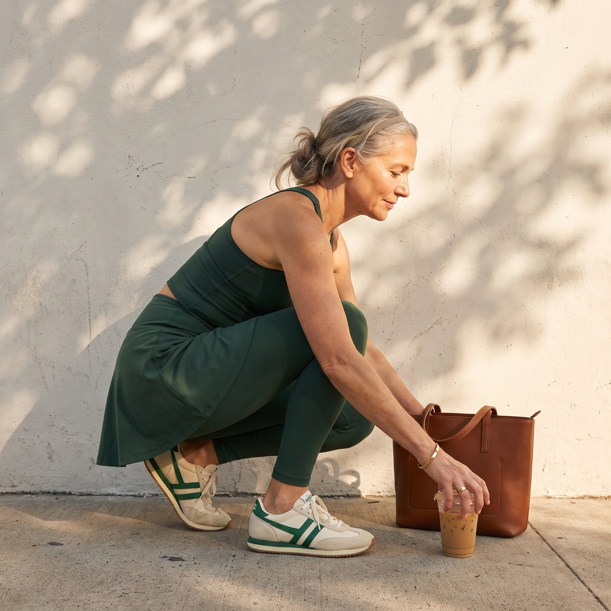 A woman in a dark green activewear set crouches on a concrete patio against a textured wall, holding an iced coffee next to a brown leather tote bag.