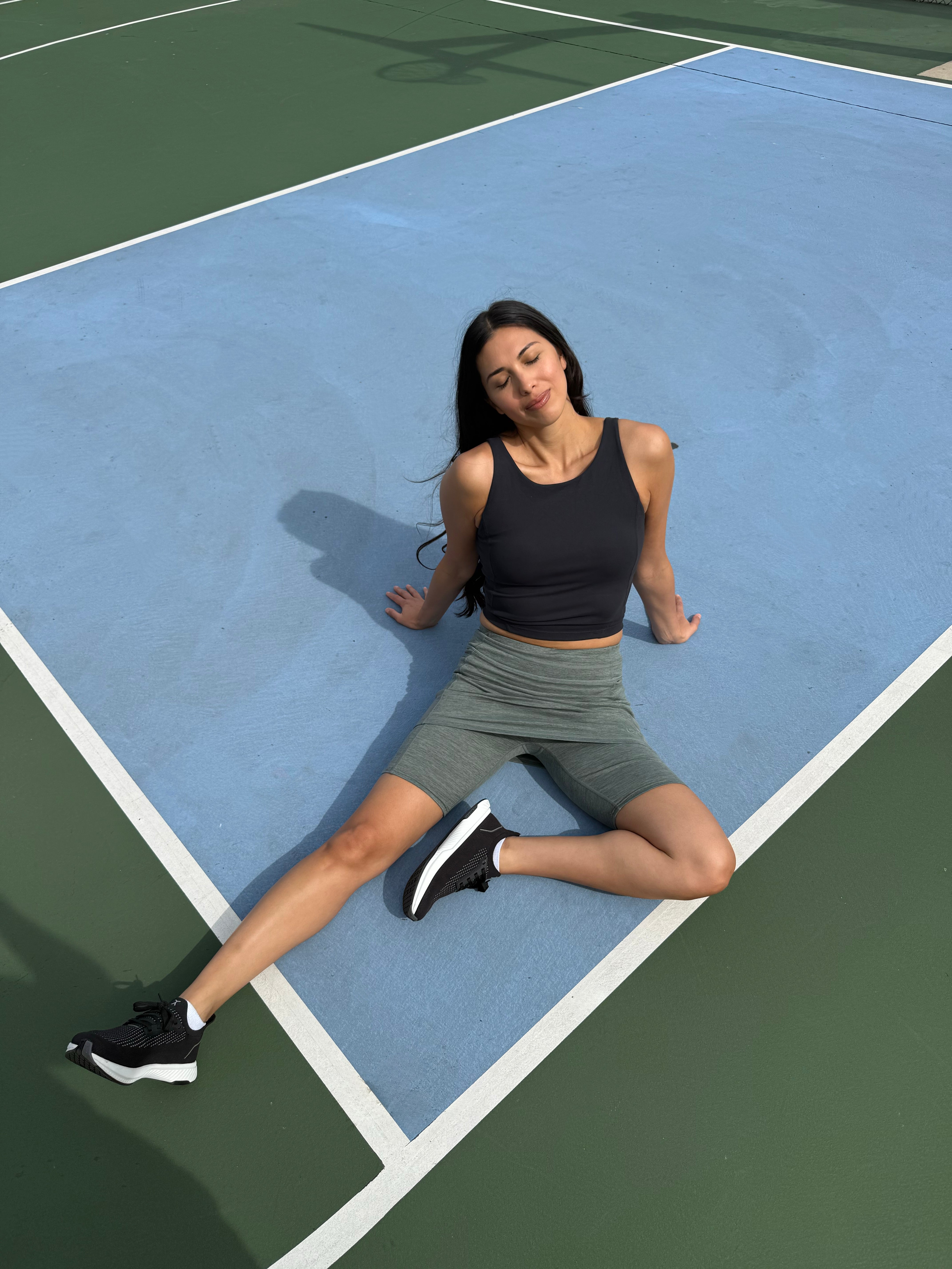 Woman stretching on a blue and green tennis court wearing the Fern Grey Heather Bike Short Skirted leggings. 