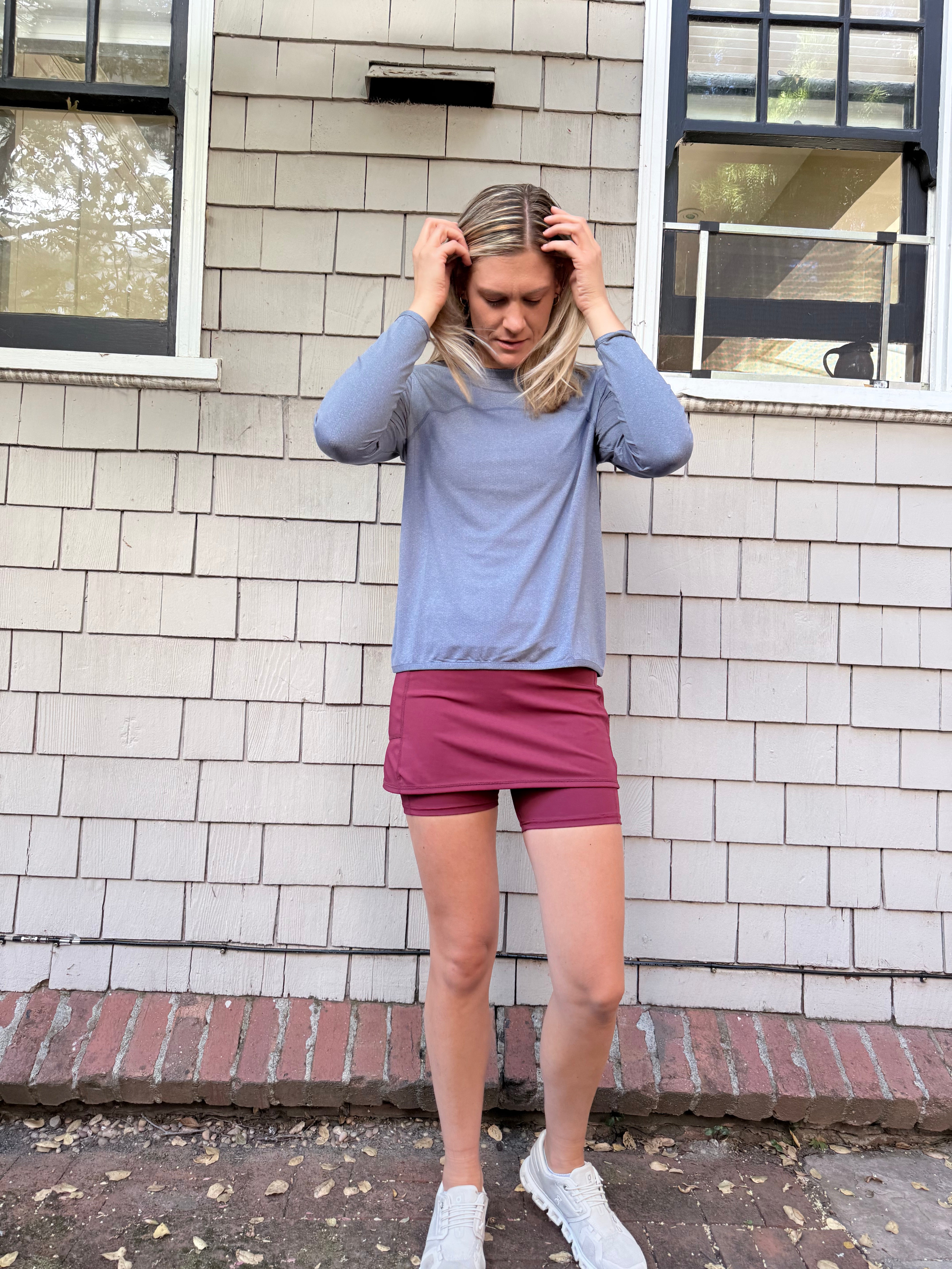 A woman stands outdoors against a white brick wall wearing a light Blue Ventura Pullover Mesh Shirt athletic top, a burgundy athletic mini skirt, and white sneakers.