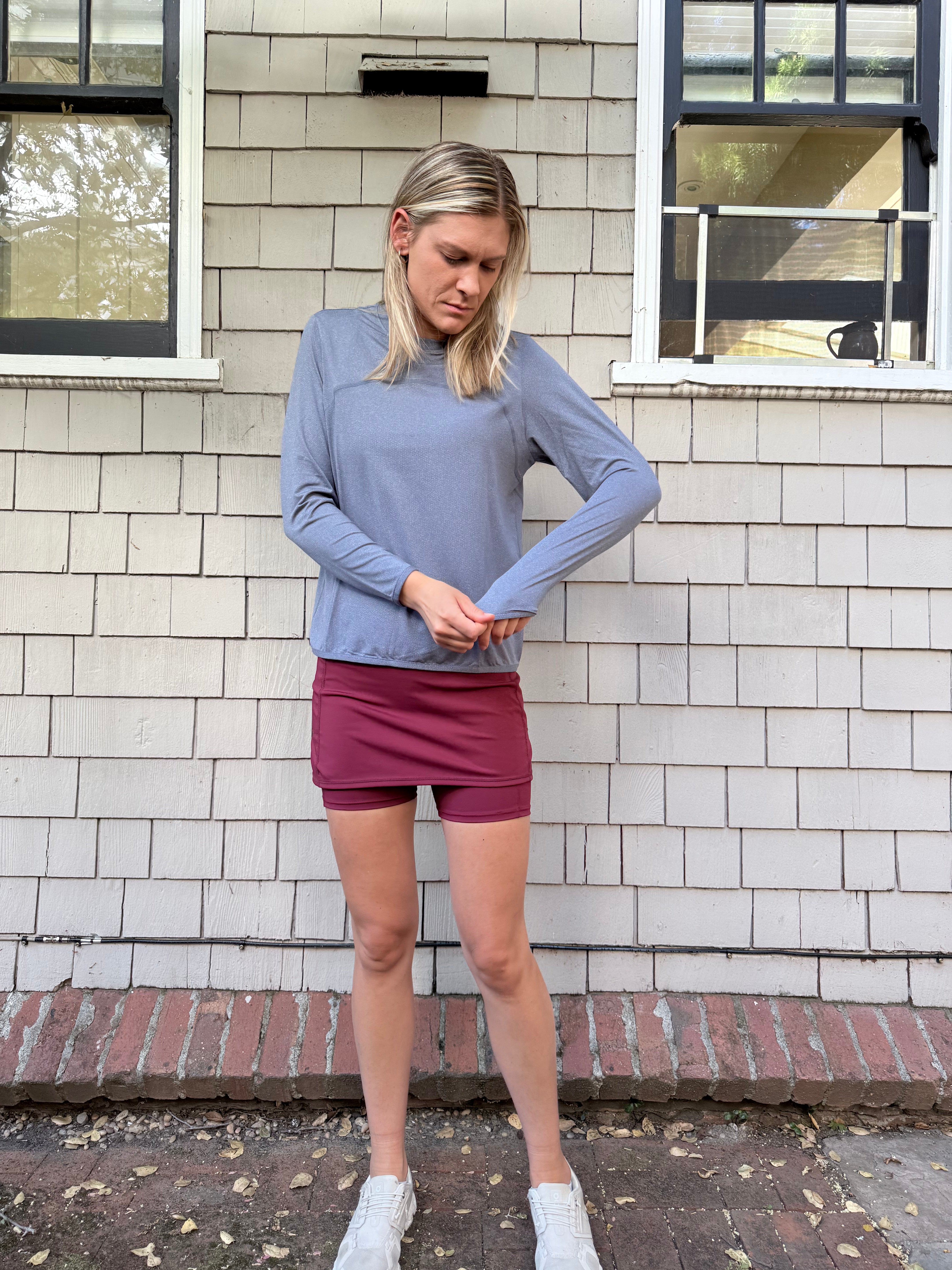 A woman stands outdoors against a white brick wall wearing a light Blue Ventura Pullover Mesh Shirt athletic top, a burgundy athletic mini skirt, and white sneakers.
