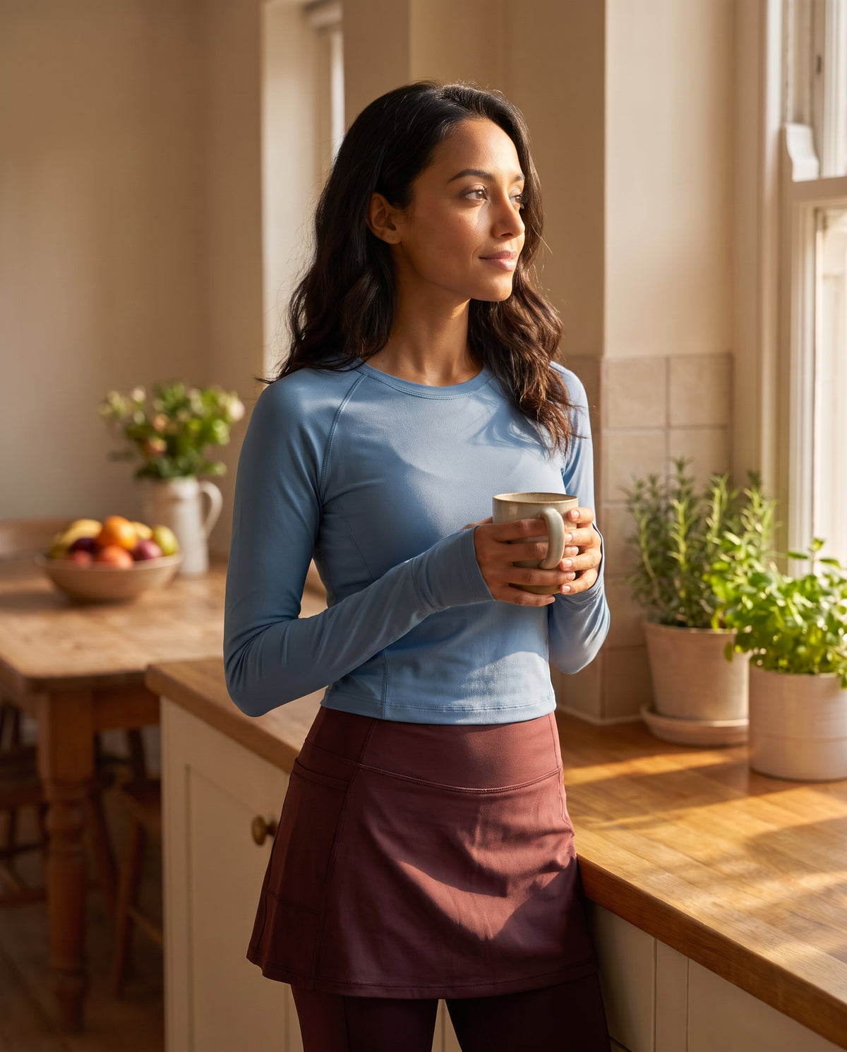 A woman stands in a sunlit kitchen wearing a light blue long-sleeve athletic top and a burgundy wrap skirt, holding a mug near a window.
