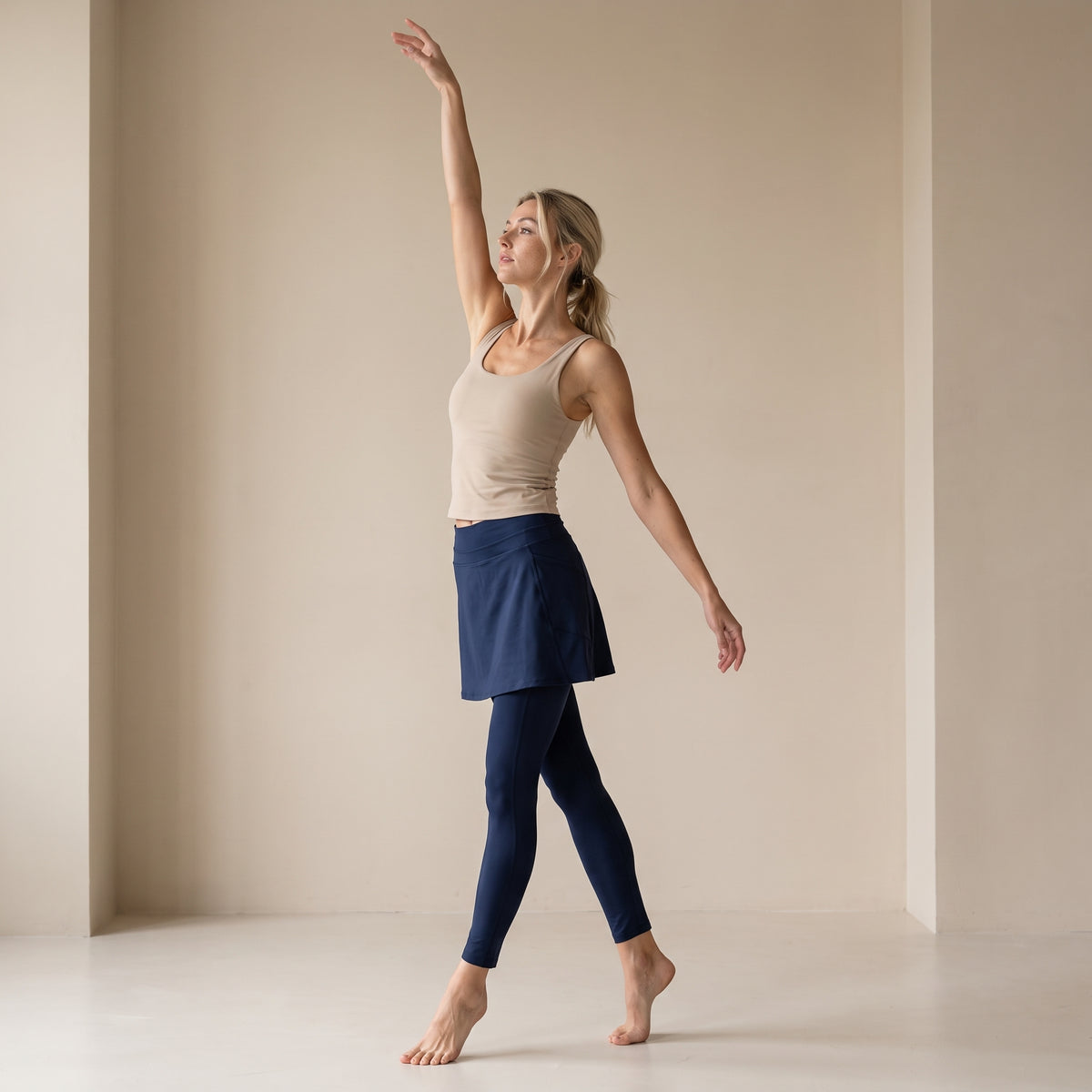 A woman in a beige tank top and navy blue leggings poses in a minimalist studio with beige walls, showcasing a navy blue athletic skirted leggings.