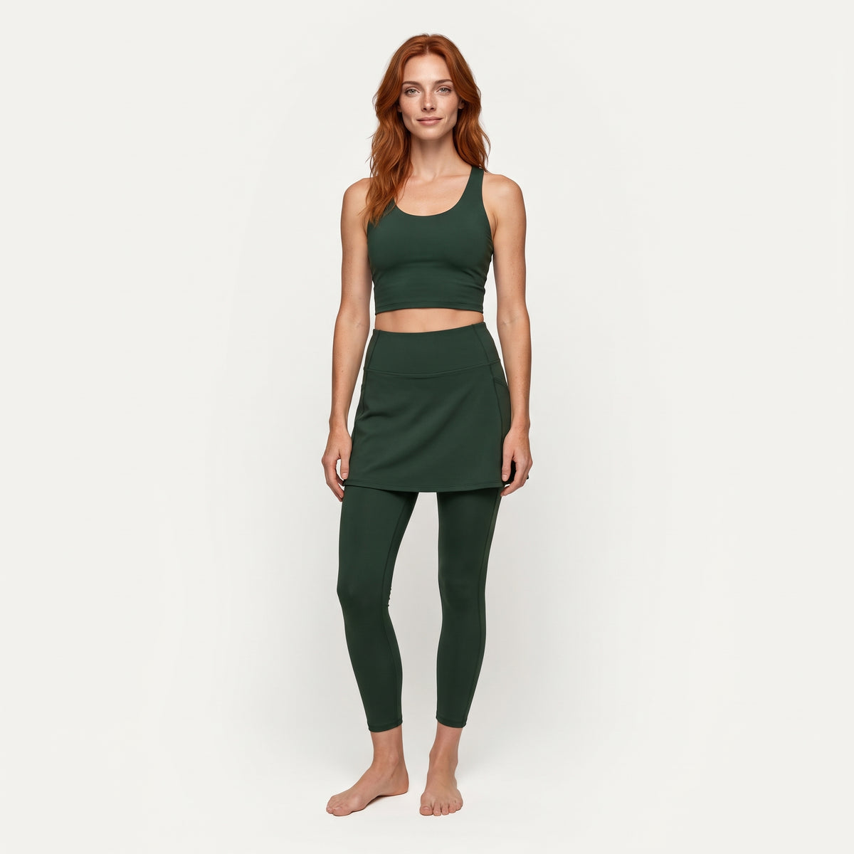 A woman modeling a dark green athletic set consisting of a scoop-neck crop top and a matching high-waisted mini skirt with leggings, shot against a solid off-white background.