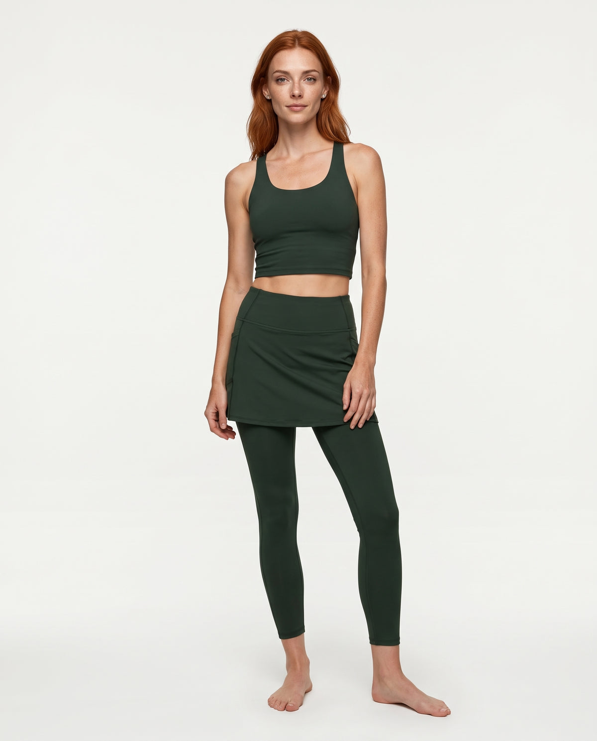 A woman modeling a dark forest green activewear set consisting of a scoop-neck crop top, a matching mini skort, and full-length leggings, shot against a plain white studio background.