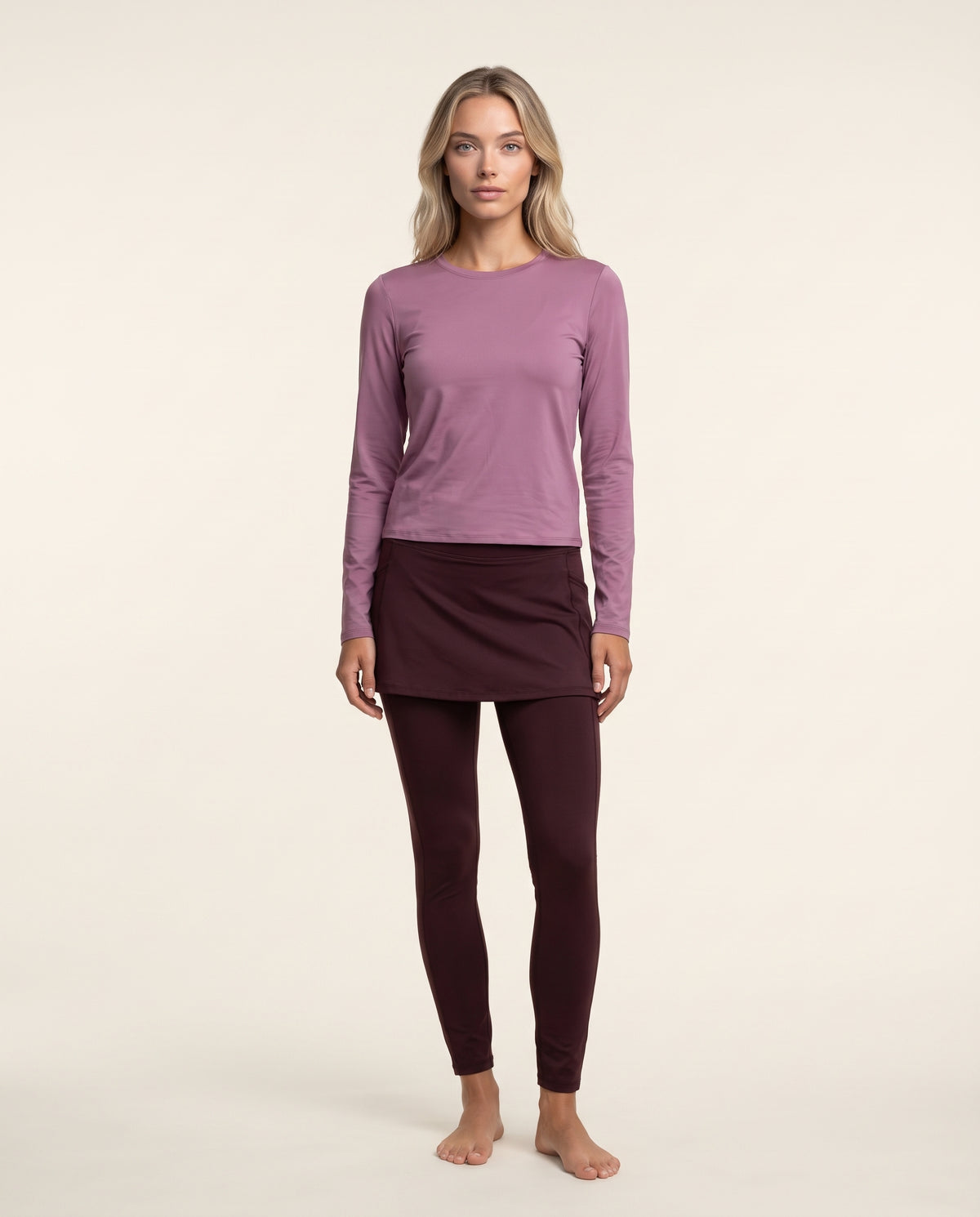 A woman modeling a two-piece lounge set consisting of a dusty purple long-sleeve top and dark burgundy leggings, standing against a solid off-white background.