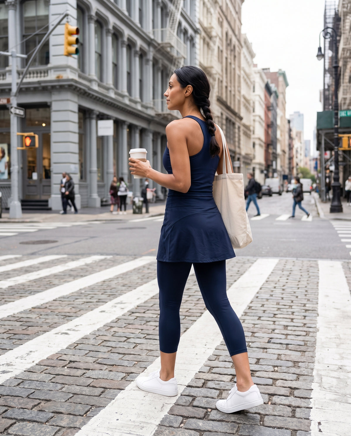A woman in a navy blue activewear set consisting of a sleeveless top and skirted leggings, walking across a city crosswalk while holding a coffee cup and carrying a beige tote bag.